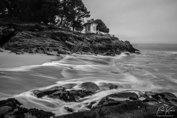 Photo en noir et blanc, au bord de la mer, présentant une petite plage de sable dans une crique bordée par des falaises de quelques mètres. Sur la plage, on voit le filé du ressac généré par une ouverture d'un tiers de seconde de l'obturateur. Au sommet de la falaise, on voit un petit cabanon de style Rococo ((?)) entre des pins maritimes.
