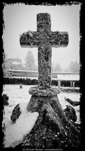 A cross on a cold winter day. Snow covers the base of the cross and the garden in the background.
