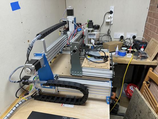 A wide-angle view of a CNC router with a spindle motor, control electronics, and a partially constructed drag chain. The bench holds various electronic components and tools, indicative of an active project in progress.
