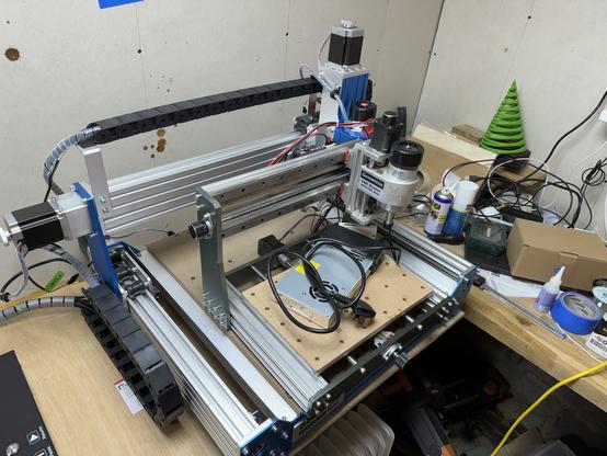 Overhead view of a CNC machine with blue stepper motors, a control board, and wiring. A spindle with a dust collection attachment is at the center, set against a backdrop of a cluttered workshop bench with tools and materials.