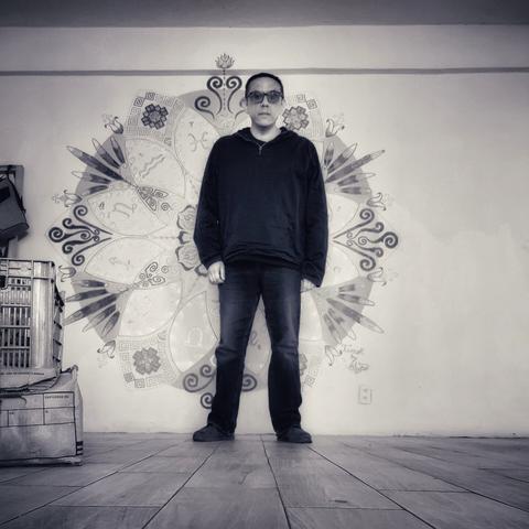 Un hombre vestido de negro está de pie en el fondo de un cuarto, ligeramente a descentrado respecto de un mandala pintado en la pared.