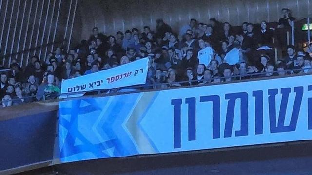 Crowd of people behind a railing with banners, one displaying Hebrew text which say “only transfer will bring peace”
