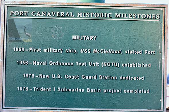 A photograph of a sign with a green background and brass-colored border and text reading, “Military 1953 – First military ship, USS McClelland, visited Port 1956 – Naval Ordnance Test Unit (NOTU) established 1976 – New U.S. Coast Guard Station Dedicated 1978 – Trident I Submarine Basin project completed