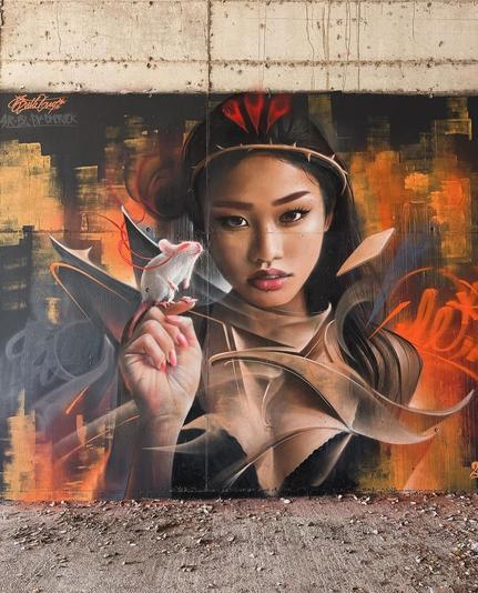 Streetartwall. The beautifully designed portrait mural of an Asian girl with a white mouse was sprayed/painted on a rough, beige concrete wall. The portrait of the beauty with long black hair, wearing a crown of thorns and a tight black outfit, is framed by a stylized city silhouette in shades of red, black and orange. The girl has a penetrating gaze, a realistically designed fine face and holds up a hand on which sits a white mouse with red eyes. Beige and gray graffiti waves circle her.