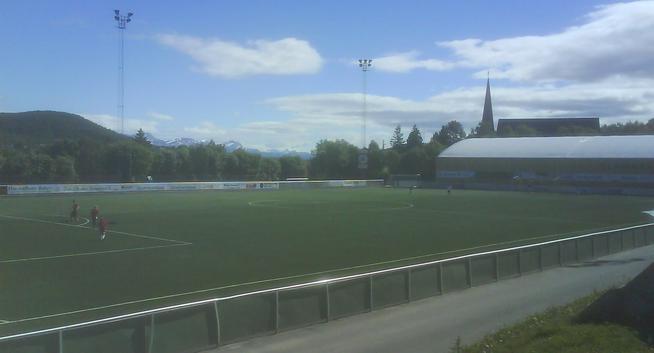 Fotografi av ein nesten tom fotballbane med ein idrettshall synleg i bakgrunnen.