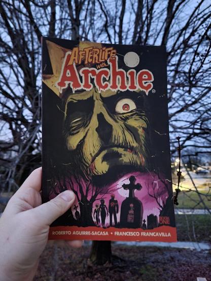 Photograph of a white person's hand holding the first volume of Afterlife With Archie in the air with a tree with no leaves seen in the background. The cover of the book has an ominous, partially decayed face of Jughead over looking various silhouettes of characters at the ground surface.