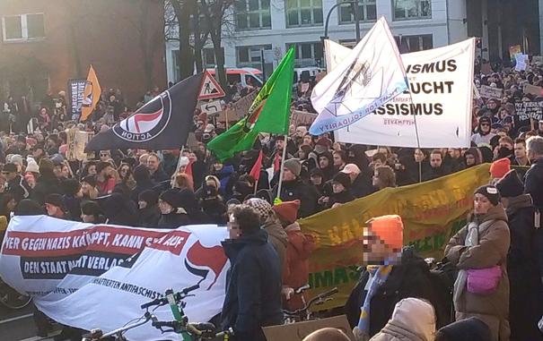 Foto des Antikapitalistischen Blocks in der Menschenmasse, fotografiert von schräg vorne. es sind Banner und Antifa-Fahnen zu sehen. Die Schrift ist schwer zu erkennen, da die Banner im Wind wehen.