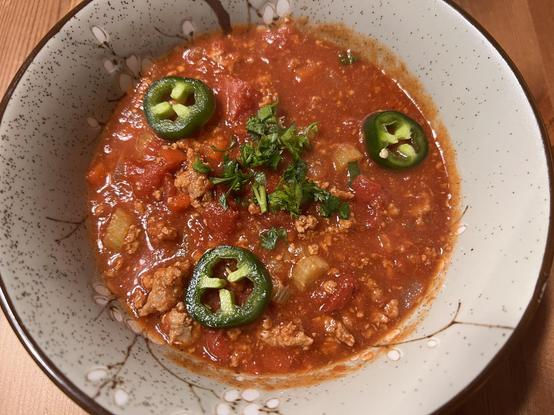 A photo of a bowl of red chili, visible are bits of ground meat (turkey), celery, and tomatoes. The chili is topped with jalapeño slices and some diced parsley.
