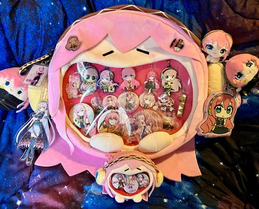 A plush Megurine Luka ita bag full of buttons and keychains. The bag resembles a stylized Luka face with the display window being her gigantic open mouth.