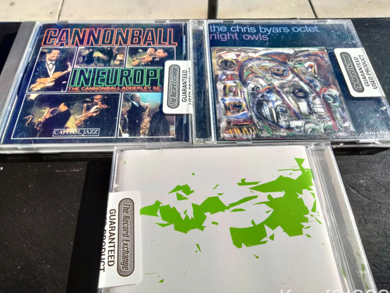 Three CDs on a black table, with a strip of concrete sidewalk visible above. Several color shots of the sextet performing; an expressionistic mishmash of a painting; mysterious green angular shapes like a Mercator projection of mysterious green continents of a white Mercator ocean world. There is a ~lot~ of ocean on top.