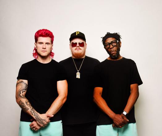 Three men staring at the camera intently

The two on the left and right are wearing black T-shirt and turquoise pants, visible to mid thigh. The man in the middle is wearing all black —  shirt and pants — we can’t see his arms

The man on the left of the photo is very light skinned White with snake-like tattoos on his right arm while his left arm has no markings. He has bright red hair

The man on the right is Black with dark, wide black glasses and hair tied back in a black bandana. His untattooed arms are crossed in front of him like the man on the left

The man in the middle is White in all black clothing with a Pittsburgh Pirates baseball cap, red-shaded sunglasses, and a necklace of perhaps Buddhist design. His arms are hidden behind the men to his left and right