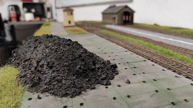 Railroad model module with platform and pile of dirt or coal on the side of a platform, behind the module microwave, coffee machine and whiteboard can be seen, destroying the realism of precisely prepared landscape