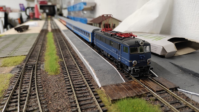 On a model railroad module, flanked by platforms on both sides, the right track features an EP08-013 (Pafawag 102E) locomotive with pantograph up sporting a distinctive blue color, dating back to approximately 2007-2009. Notably, it lacks the Intercity logo and has transitioned from orange. Following the locomotive are two light blue couchette wagons (110Ac) and likely a 112ag passenger carriage.