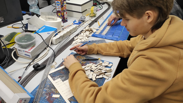 Boy with light brown hoodie cutting out plywood parts from lasercut sheet, to the left another person is laying them on masking tape probably for coating them afterwards with paint, track going through the table, some containers with parts or tools, there is also a WOŚP (great orchestra of christmas charity) collection box standing in a background