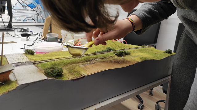 Railway model module with one track going across it, with pedestrian railway crossing made of concrete slabs between grassy field with bushes, a person preparing the module sprinkles the surface with grass using a fine sieve