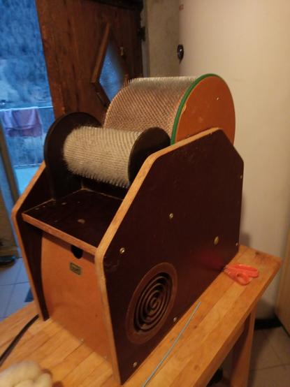 Das elektrische Kardiergerät.
Zwei unterschiedliche grosse benadelte Walzen sind auf einem Sperrholzkasten montiert. Es ist ein Gerät der Firma Louët aus den 80er Jahren