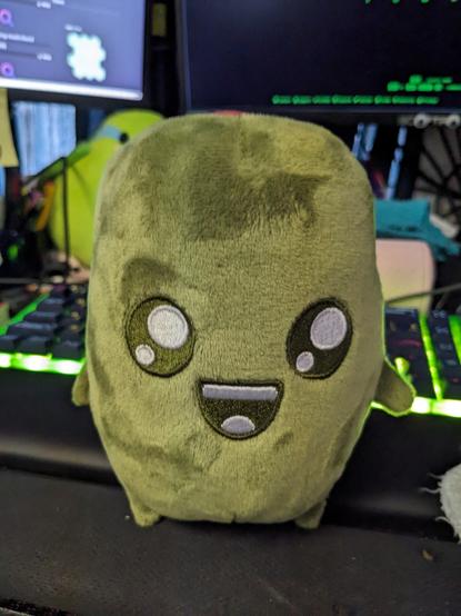 olive plushie that is smiling in front of a keyboard on a cluttered desk