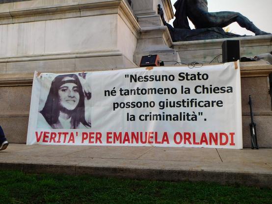 a big banner  using the same picture ,   next to the text :    "Nessuno Stato  né tantomeno la Chiesa  possono giustificare  la criminalità  -  VERITÀ PER EMANUELA ORLANDI"    ('No State,  nor even the Church,  can justify crime  - TRUTH FOR EMANUELA ORLANDI')