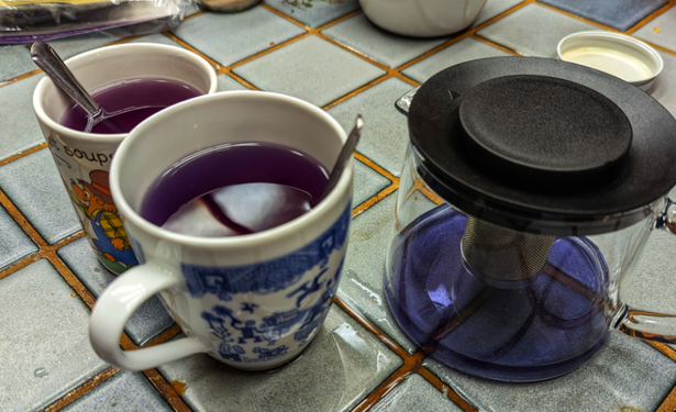 Two mugs of purple tea sit beside a glass teapot filled with blue tea.