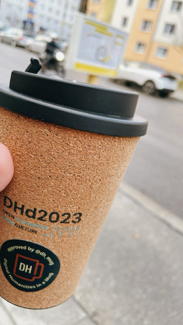 Ein Kork-Kaffeebecher in einer Hand. Im Hintergrund eine Bushaltestelle