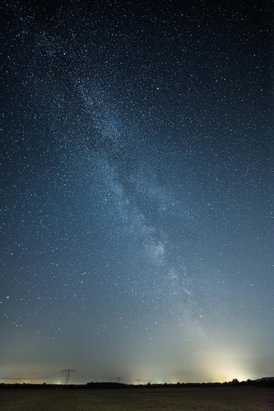 EN: An upright photo of the Milky Way. Scattered villages can be seen on the bottom of the picture. From the villages shines golden light pollution to the sky.

DE: Ein Hochkantfoto der Milchstraße. Am unteren Bildrand sind vereinzelte Dörfer zu sehen, die bis zu einer gewissen Höhe den Himmel goldgelb einfärben.