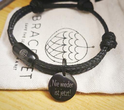 Ein schwarzes Armband aus alten Fischernetzen, so genannten Geisternetzen. Am Armband befindet sich ein Anhänger auf dem steht "Nie wieder ist jetzt"