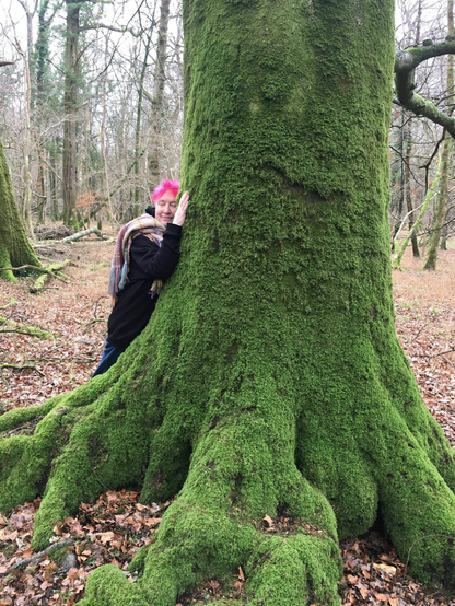 A woman with pink hair hugs the trunk of a huge old beech tree covered in bright green moss