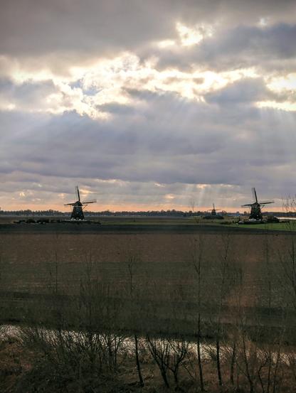 Wide fields with 3 wind mills on them and the sun breaking through the clouds