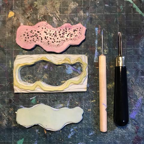 Three small Lino blocks with an awl and a carving knife on a cutting mat. The upper and lowest blocks are irregular organic shapes and the middle block looks like the exterior of such shapes. The upper block has many tiny round holes in it.