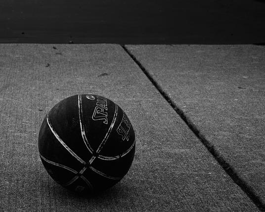 ourt, its once bold lines faded from use and time. The court is deserted, with no players in sight, giving the scene an air of abandonment. The concrete ground is marked by cracks and the subtle textures of wear, hinting at the many games played here. The basketball, branded 'Spalding,' sits slightly off-center in the frame, drawing the eye as the main subject. The absence of color in the image accentuates the starkness of the scene and the ball's solitary presence. The overcast sky suggests a chilly season, either autumn or winter, contributing to the feeling of loneliness and the anticipation of activity's return. Despite the stillness, there's a latent energy, as if the ball is poised and waiting for the next game to commence