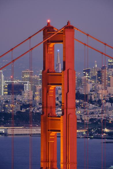 Close-up view of a pillar of the Golden Gate Bridge. A view on the Transamerica Pyramid is lined up through one of the openings. Blue hour.