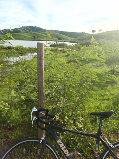 Foto de uma bicicleta speed preta escorada em um toco de cerca de uma área rural com um lago ao fundo e, após ele, vários morros desmatados para pastagem.