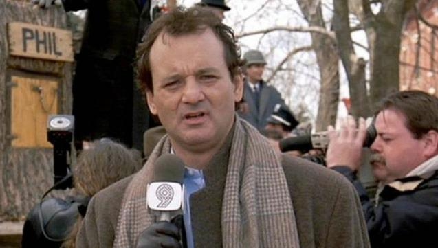 Bill Murray, here a young man, though with a sour expression, is holding a Channel 9 news mic in front of a tree stump. He’s wearing a coat, scarf, and gloves, and is reporting on a busy event.