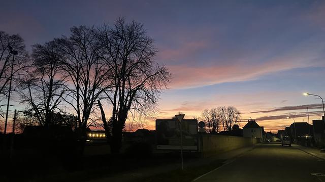 Eine Straße in der Abenddämmerung mit silhouettierten Bäumen vor einem bunten Himmel. Die Straßenlaternen sind eingeschaltet, und auf der linken Seite ist eine Werbetafel zu sehen.