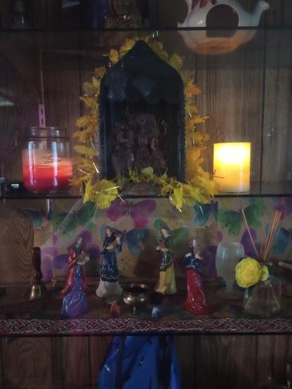 On the top shelf is black shrine box with yellow flowers draped over it. It frames a carving of the Three Brigids. Brigid of Poetry faces forward, hands raised, as though giving an oration. Brigid of Healing faces left, holding a baby. Brigid of Smithcraft is facing right and pounding on an anvil.
Two large lit candles are on either side of the box. on the shelf below are ceramic statues of 4 female minstrels playing music around a cauldron that is surrounded by rocks of various colors.
