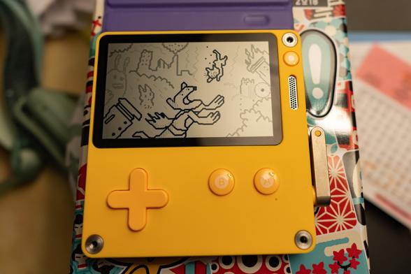 Une console Playdate avec le programme "Playmaker", avec un dessin d'un chaton sauvé par un super-héros chien dans une ville en flammes. La manivelle n'est pas active.