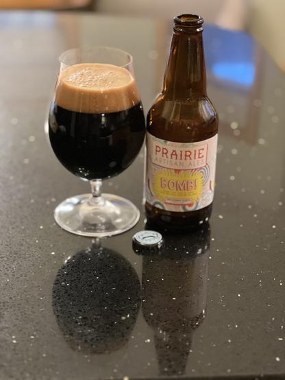 A bottle of Prairie Ales BOMB! Beer. Deep black with a dark caramel head. The label is white with red print and accents in yellow blue and red and brown