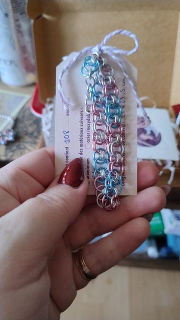 Le bracelet en mailles, aux couleurs du drapeau trans, bleu pastel+rose pastel+blanc+rose pastel+bleu pastel, accroché à sa propre carte de présentation: 108 anneaux en aluminium