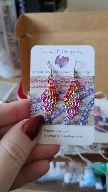 Les boucles d'oreilles aux couleurs du drapeau lesbien, orange foncé+orange clair+blanc+rose clair+rose foncé, accrochées à leur petite carte.