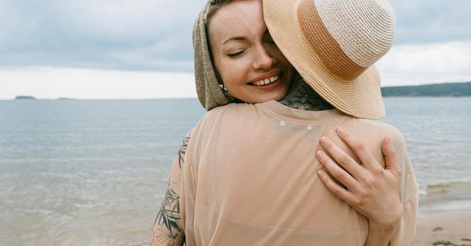 Image of two women hugging each other on a beach with water in the background.