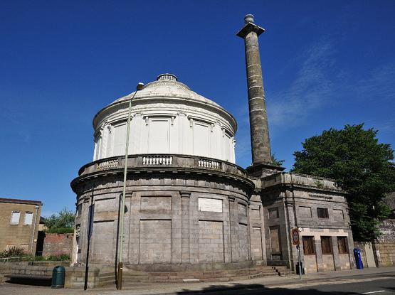 The Fergusson Gallery, housed in Perth's old waterworks building. The image shows a circular stone building with a white rotunda on top of it. To its right a tall stone chimney sits on top of a square stone wing of the building. The sky is blue.