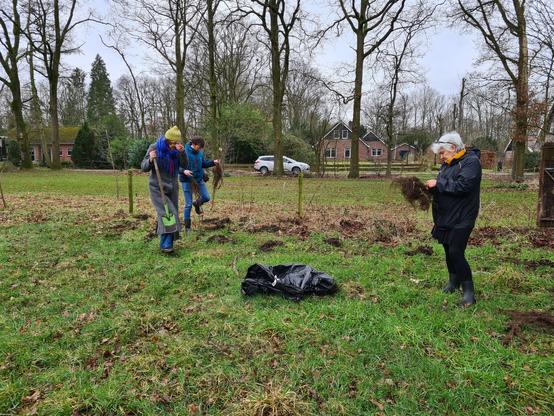 Drie mensen bij de erfgrens in een ruig weiland met plantgaten, één persoon met een spade, 2 mensen met plantmateriaal. Grijs weer.