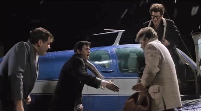 Four young men in 50s clothing and jackets hurry to put their bags and belongings on a small blue plane