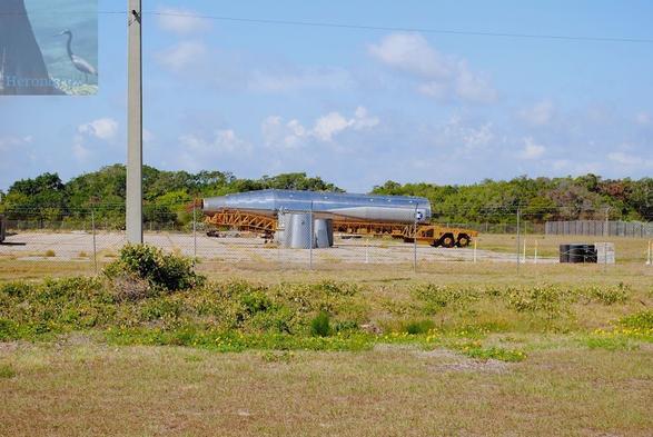 An outdoor landscape photograph of an old, shiny metal rocket body on a horizontal carrier with several rocket parts nearby. There is an open grass field starting at the bottom of the frame and extending to a chain-link fence around the concrete pad on which the rocket parts rest.