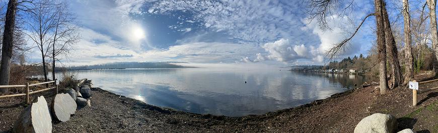A panoramic photo of Lake Washington taken from Log Boom Park in Kenmore showing sunny skies with various cloud types and foggy trees reflected in the water.