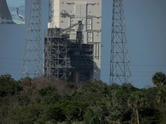 A photograph of part of Space Launch Complex 37B. At the bottom of the frame are green trees and scrub land, above which power lines are strung. Behind them and reaching past the top of the frame is a white tower. The lower section is open and a single orange cylinder can be seen inside, which is a Delta 4 rocket core. The upper part of the building is closed in by doors that slide sideways to open. There are two tall truss towers on either side of the white tower, and a truss building just on its left side with connections reaching to the rocket inside the white tower.