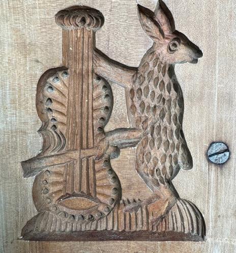 A wooden board from Germany for forming cookies. Carving of a standing hare with arms holding a standing stringed instrument (cello). The instrument is on the left and the hare is on the right, with its head turned to the right also. There is a screw on the right where the board was repaired from a large split.
