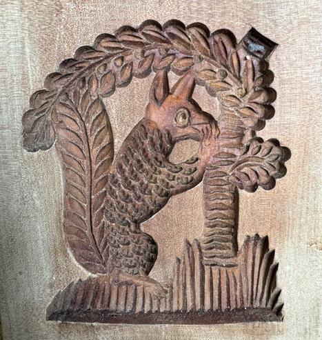 A wooden board from Germany for forming cookies. Carving of A squirrel on the left, facing right. Carving on the right has horizontal lines and leaves, including a swooped leaf going above and over the squirrel.