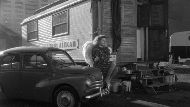 Solveig Dommartin, a beautiful white French woman with curly blonde hair, sits on the hood of an old car. She is wearing angel wings and hugs a large jacket to her body.