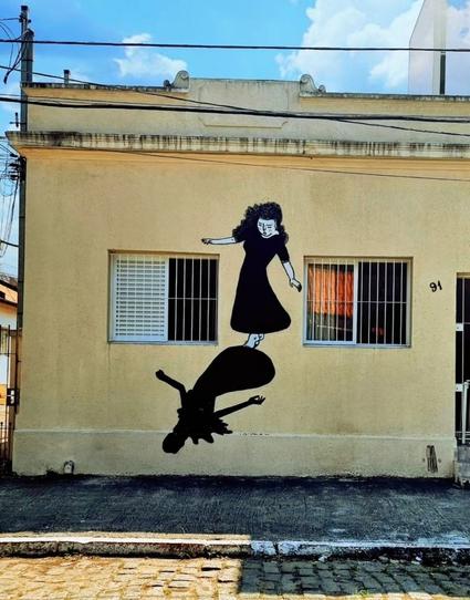 Streetartwall. A black and white mural of a dancing girl with her shadow was sprayed/painted on the yellow wall of a simple flat building. The house has two barred windows and a piece of the door can be seen. The mural with the girl is drawn in a minimalist style. She has long curly hair and is wearing a black dress. She appears to be jumping or dancing, holding her arms out to either side and looking down at her feet. Her shadow can be seen there. 
The environment in the photo is a blue sky with some clouds.
Info: Alex Senna is a very well-known Brazilian artist known for his minimalist and expressive figures that often convey feelings of love, loneliness and freedom.